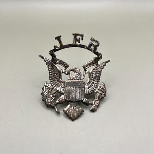 May include: Silver-toned metal pin with an eagle and shield design. The eagle is holding a banner with the letters "JFR" on it.