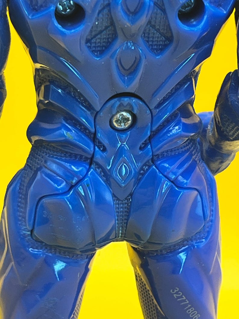 May include: Close-up of a blue action figure's back, with detailed molded features. The figure has a glossy finish and a textured design on the back and legs. A silver screw is visible at the figure's center.