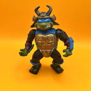 May include: A Teenage Mutant Ninja Turtle action figure, featuring a green turtle head with a blue mask and a samurai helmet. The figure has a gold chest plate, black arms and legs, and blue accents. The figure is posed against a bright orange background.