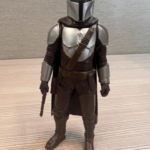 May include: A detailed action figure of The Mandalorian, featuring a silver helmet and armor, a dark brown outfit, and a blaster. The figure stands upright, showcasing the character's iconic design from the Star Wars series.