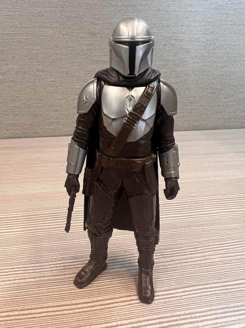 May include: A detailed action figure of The Mandalorian, featuring a silver helmet and armor, a dark brown outfit, and a blaster. The figure stands upright, showcasing the character's iconic design from the Star Wars series.
