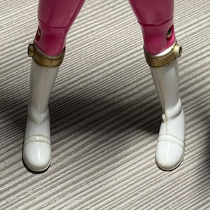 May include: A close-up of a toy figure's legs. The figure wears bright pink pants and white boots with gold-colored accents around the ankles. The boots have a smooth, glossy finish.