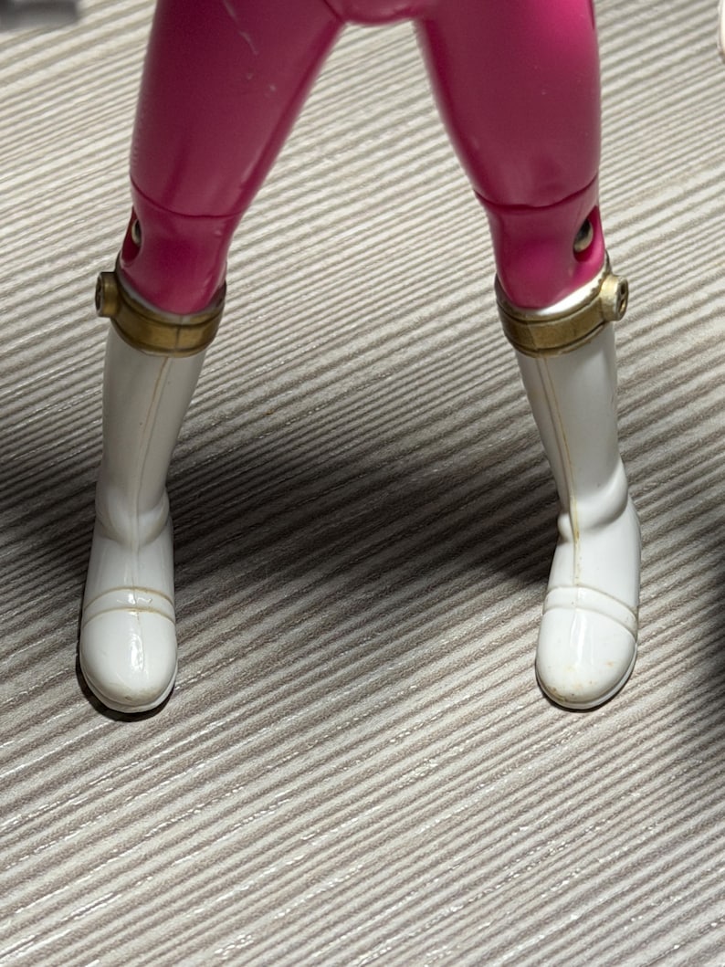 May include: A close-up of a toy figure's legs. The figure wears bright pink pants and white boots with gold-colored accents around the ankles. The boots have a smooth, glossy finish.