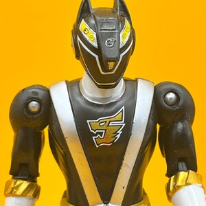 以下が含まれることがあります： 黒いアクションフィギュア。銀と金の装飾が施されています。ヘルメットには黄色のアクセントがあり、胸には狼の頭のエンブレムが付いています。腕と脚は黒で、手首とベルトには金色の装飾が施されています。