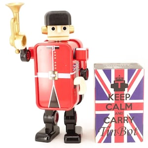 May include: A red and black wind-up tin robot toy dressed as a British guard with a gold trumpet. The robot is standing next to a box with a Union Jack design and the text "Keep Calm and Carry Tin Bot".