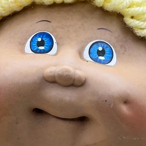 May include: Close-up of a vintage doll's face with bright blue eyes and a small, smiling mouth. The doll has a light skin tone and a yellow yarn hairstyle. The doll's face is a classic toy.