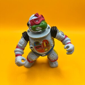 May include: A Teenage Mutant Ninja Turtle action figure in a white and gray spacesuit with red and blue accents. The figure has a green face, a red mask, and black shoulder pads. The figure is posed on a yellow background.