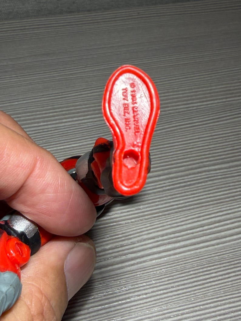 May include: Close-up of a red plastic action figure foot. The foot is stamped with the text "&copy; 1993 MARVEL" and "TOY BIZ, INC." The figure is red, black, and gray.