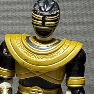 May include: A Power Rangers action figure with a black and gold helmet and armour. The figure has a gold chest plate, belt, and armbands. The figure's arms and legs are black and white.