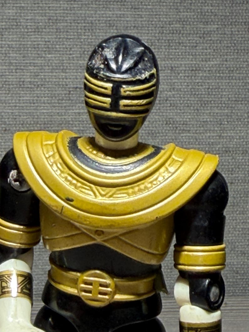 May include: A Power Rangers action figure with a black and gold helmet and armour. The figure has a gold chest plate, belt, and armbands. The figure's arms and legs are black and white.