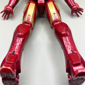 May include: Red and gold action figure of Iron Man with articulated limbs. The figure is posed with its legs spread apart and its arms outstretched. The figure is made of plastic and has a detailed paint job.