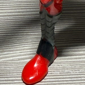 May include: Close-up of a red and black action figure leg. The foot is a solid, glossy red, while the leg has a segmented, gray design with black accents. The figure appears to be a superhero or sci-fi character.