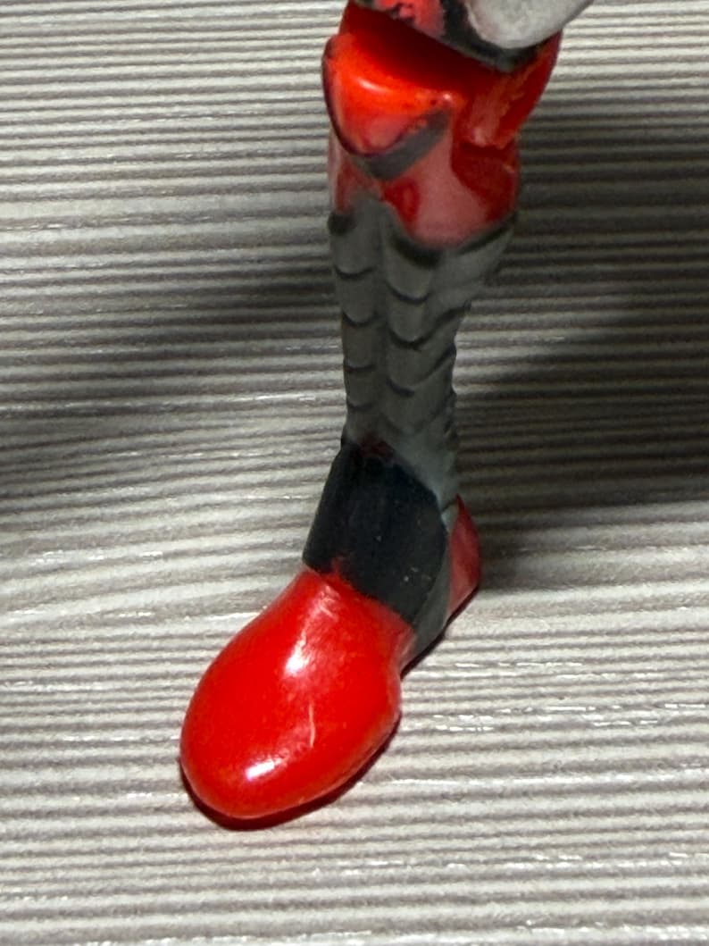 May include: Close-up of a red and black action figure leg. The foot is a solid, glossy red, while the leg has a segmented, gray design with black accents. The figure appears to be a superhero or sci-fi character.