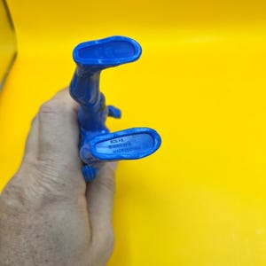 May include: A blue plastic action figure, seen from the feet up, against a yellow background. The figure's feet have text that reads "SCG P.R. BANDAI 2018 MADE IN CHINA". The figure is held by a hand.