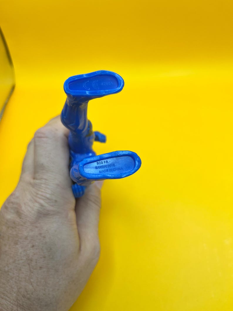 May include: A blue plastic action figure, seen from the feet up, against a yellow background. The figure's feet have text that reads "SCG P.R. BANDAI 2018 MADE IN CHINA". The figure is held by a hand.
