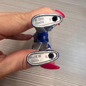 May include: A blue and silver action figure, held by a person. The figure's feet are visible, with the text "2000 BANDAI" and "MADE IN CHINA" printed on the soles. The figure is a collectible toy.
