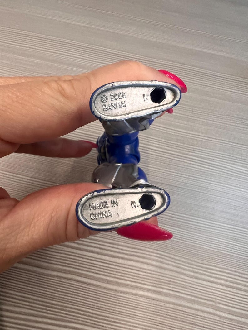 May include: A blue and silver action figure, held by a person. The figure's feet are visible, with the text "2000 BANDAI" and "MADE IN CHINA" printed on the soles. The figure is a collectible toy.