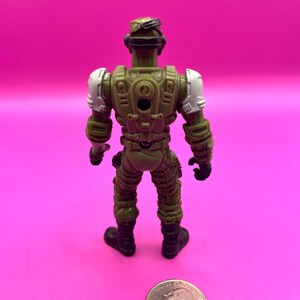 K&ouml;nnte beinhalten: Eine gr&uuml;ne und beige Actionfigur eines Soldaten, von hinten gesehen, mit wei&szlig;en Schulterpolstern und schwarzen Stiefeln. Eine M&uuml;nze liegt neben der Figur zum Gr&ouml;&szlig;envergleich. Die Figur befindet sich auf einem rosa Hintergrund.