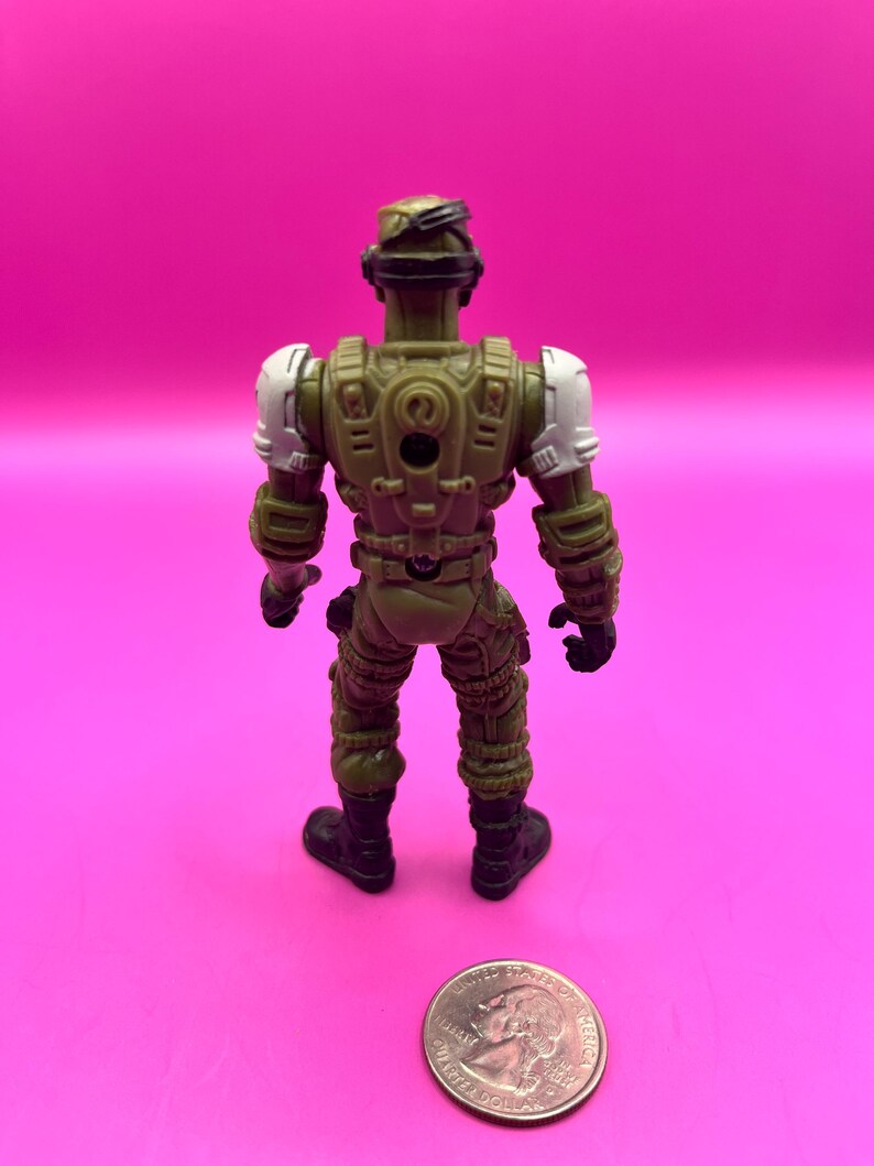 K&ouml;nnte beinhalten: Eine gr&uuml;ne und beige Actionfigur eines Soldaten, von hinten gesehen, mit wei&szlig;en Schulterpolstern und schwarzen Stiefeln. Eine M&uuml;nze liegt neben der Figur zum Gr&ouml;&szlig;envergleich. Die Figur befindet sich auf einem rosa Hintergrund.