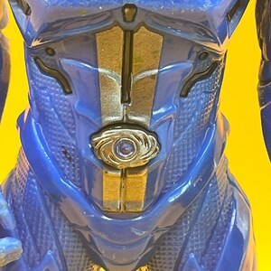 May include: Close-up of a blue action figure's torso. The figure has a textured, patterned design with gold and silver accents. A circular emblem is centered on the chest.