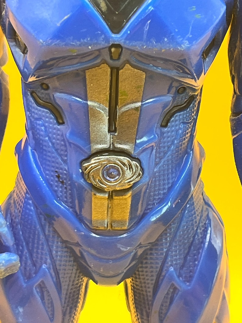May include: Close-up of a blue action figure's torso. The figure has a textured, patterned design with gold and silver accents. A circular emblem is centered on the chest.