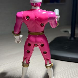 May include: A pink action figure of a superhero, viewed from the back. The figure has a white helmet with a cross design, white boots, and gold accents. It is holding a silver weapon in its right hand.