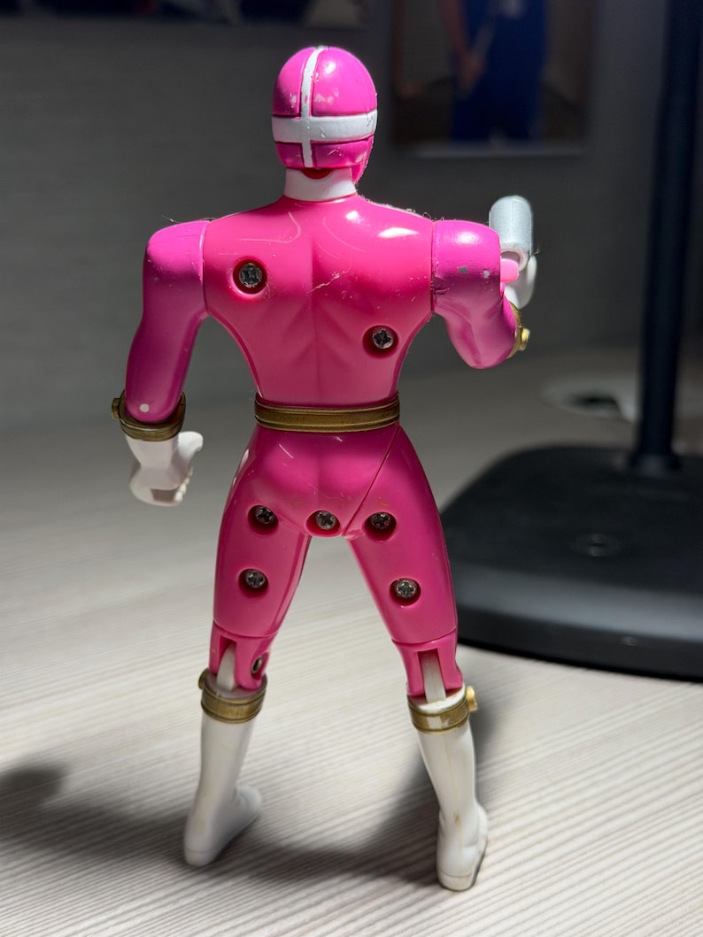 May include: A pink action figure of a superhero, viewed from the back. The figure has a white helmet with a cross design, white boots, and gold accents. It is holding a silver weapon in its right hand.