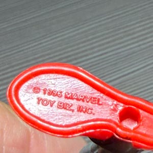 May include: Close-up of a red plastic toy base with the text "&copy; 1996 MARVEL TOY BIZ, INC." The base has a small hole and is connected to a figure with red and black details. The toy is likely a collectible item.