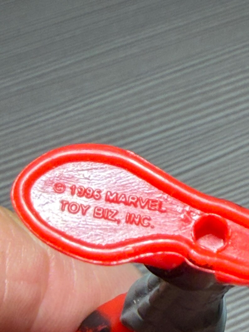 May include: Close-up of a red plastic toy base with the text "&copy; 1996 MARVEL TOY BIZ, INC." The base has a small hole and is connected to a figure with red and black details. The toy is likely a collectible item.