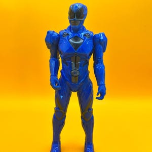 May include: A blue Power Ranger action figure, standing against a yellow backdrop. The figure has a helmet with a black visor, and a black and silver design on the chest. The figure is wearing a blue suit with armour details.