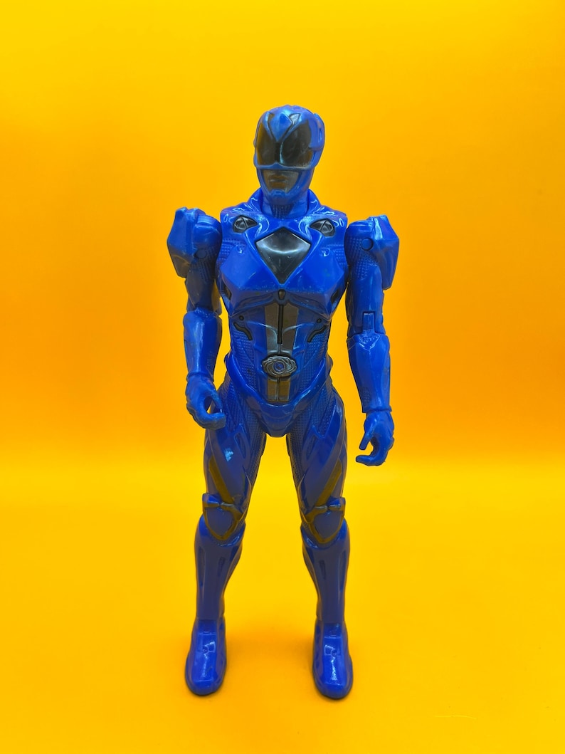 May include: A blue Power Ranger action figure, standing against a yellow backdrop. The figure has a helmet with a black visor, and a black and silver design on the chest. The figure is wearing a blue suit with armour details.