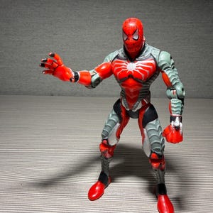 May include: A red, gray, and black Spider-Man action figure in a dynamic pose. The figure has a red head and chest with a white spider emblem. Gray armor covers the arms and legs, with red accents on the hands and feet.