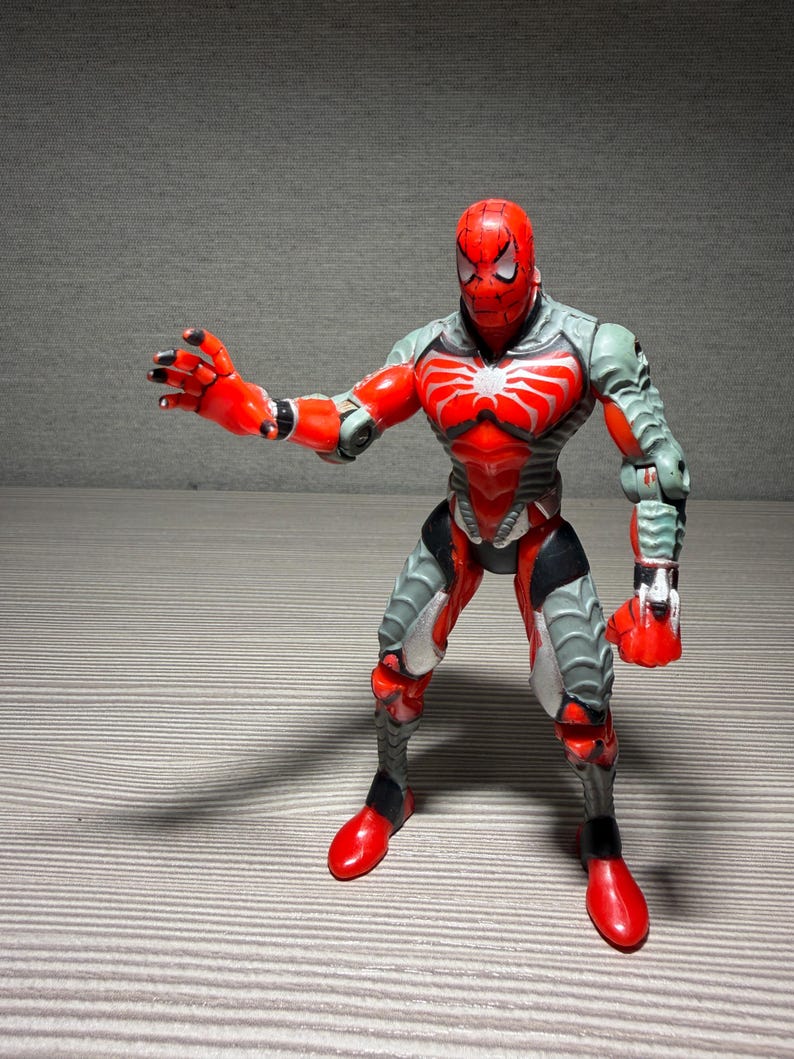 May include: A red, gray, and black Spider-Man action figure in a dynamic pose. The figure has a red head and chest with a white spider emblem. Gray armor covers the arms and legs, with red accents on the hands and feet.