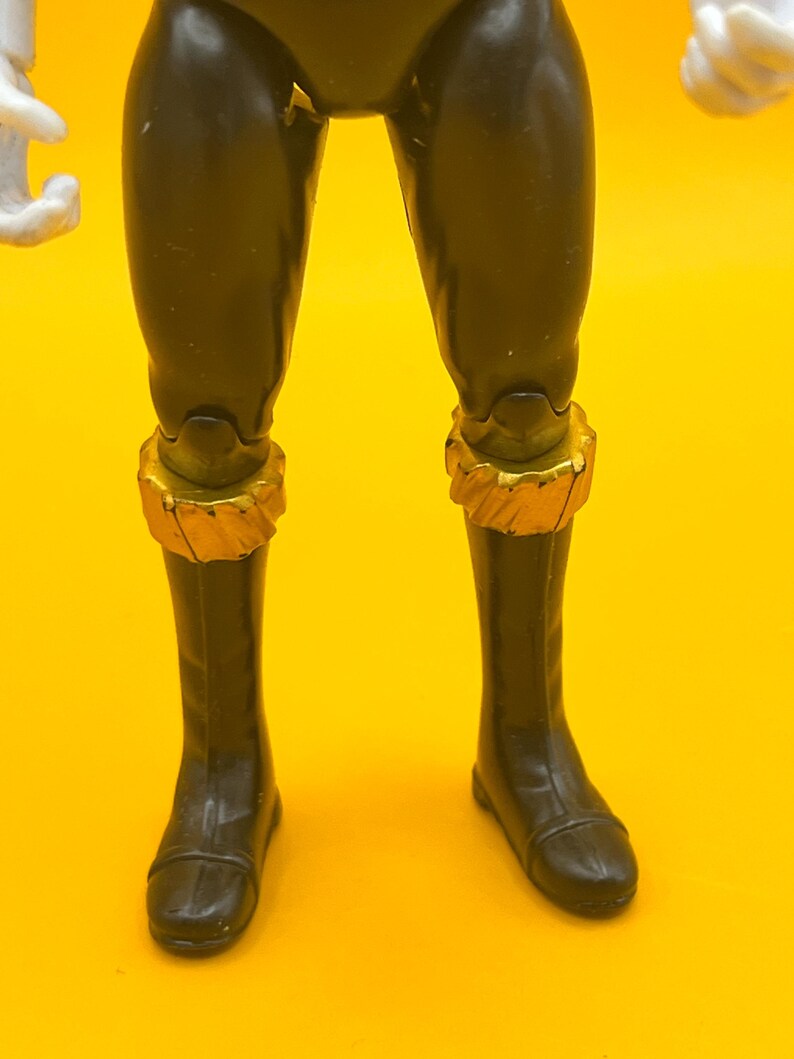 以下が含まれることがあります： 黒いアクションフィギュアの脚とブーツのクローズアップ。フィギュアは足首の周りに金色のアクセントがあります。背景は無地の黄色です。