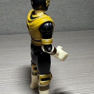 May include: A Power Rangers action figure seen from the side. The figure is predominantly black and yellow, with white gloves and boots. The helmet is black with yellow detailing. The figure is standing on a grey surface.