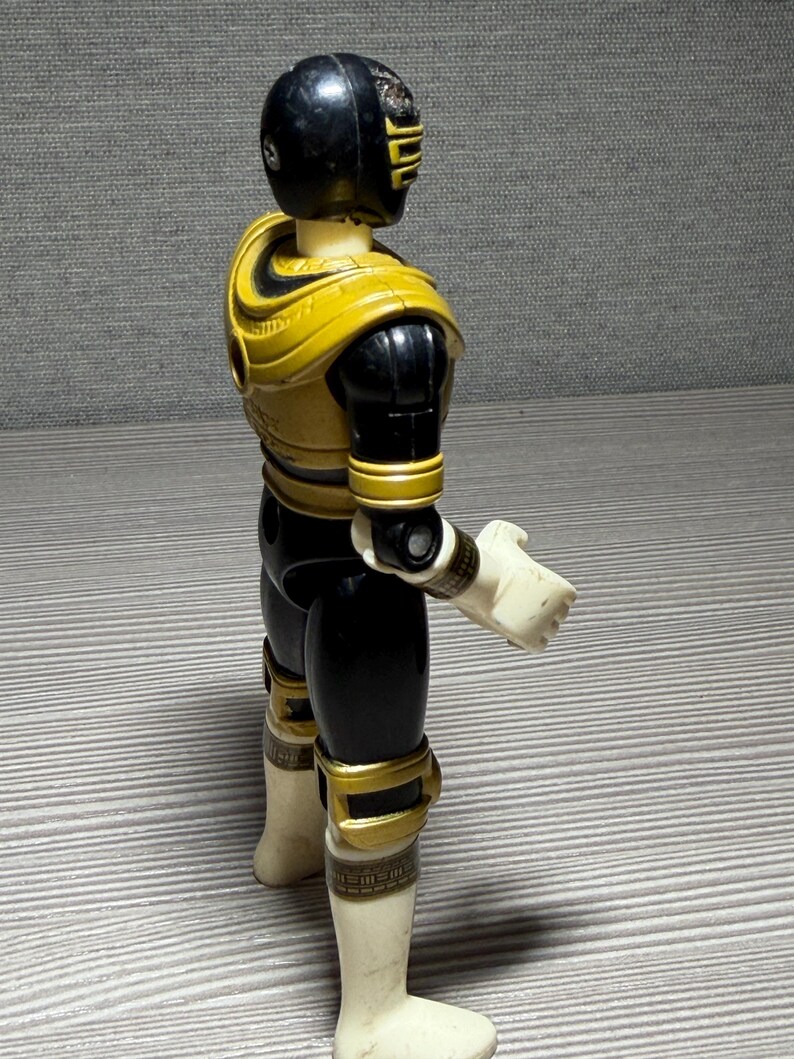 May include: A Power Rangers action figure seen from the side. The figure is predominantly black and yellow, with white gloves and boots. The helmet is black with yellow detailing. The figure is standing on a grey surface.