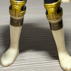 May include: Close-up of a toy figure's legs. The figure has black legs with gold knee pads and cream-coloured lower legs and feet. The knee pads have a decorative design. The figure is slightly worn.