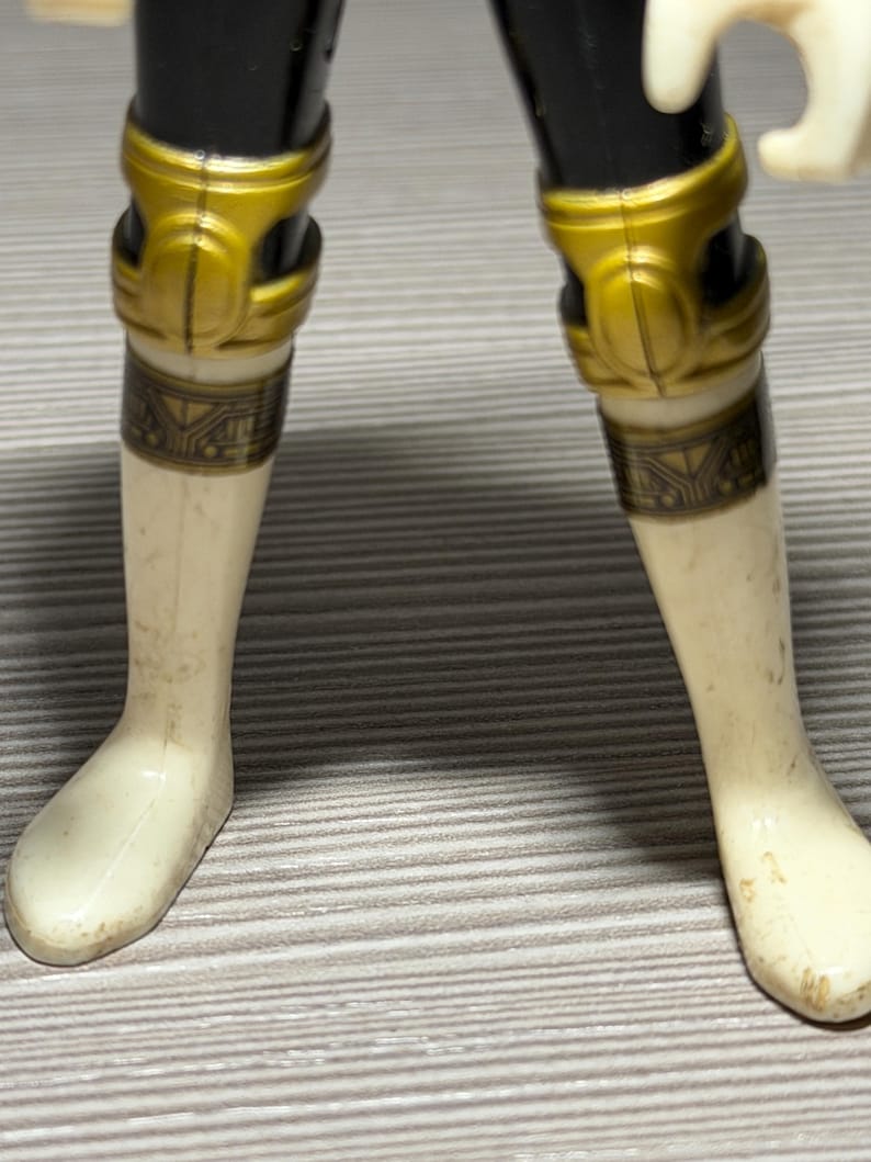 May include: Close-up of a toy figure's legs. The figure has black legs with gold knee pads and cream-coloured lower legs and feet. The knee pads have a decorative design. The figure is slightly worn.