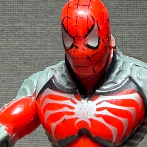 May include: A close-up of a Spider-Man action figure. The figure has a red head and torso with a white spider emblem. The figure's shoulders and arms are red, and the upper body is gray with black accents. The eyes are white with black outlines.