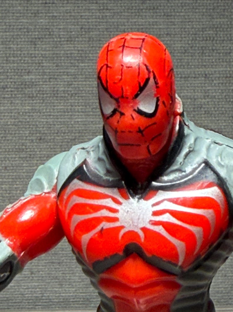 May include: A close-up of a Spider-Man action figure. The figure has a red head and torso with a white spider emblem. The figure's shoulders and arms are red, and the upper body is gray with black accents. The eyes are white with black outlines.