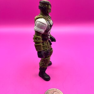 K&ouml;nnte beinhalten: Eine gr&uuml;ne und beige Actionfigur eines Soldaten mit wei&szlig;en Schulterpolstern und schwarzen Stiefeln steht auf einer rosa Oberfl&auml;che. Die Figur befindet sich neben einer M&uuml;nze. Die Figur tr&auml;gt einen Helm und eine R&uuml;stung.