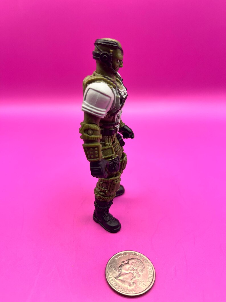K&ouml;nnte beinhalten: Eine gr&uuml;ne und beige Actionfigur eines Soldaten mit wei&szlig;en Schulterpolstern und schwarzen Stiefeln steht auf einer rosa Oberfl&auml;che. Die Figur befindet sich neben einer M&uuml;nze. Die Figur tr&auml;gt einen Helm und eine R&uuml;stung.
