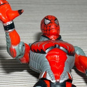 May include: A red and gray Spider-Man action figure. The figure has a red head with silver eyes and a black web design. The body is gray with red accents. The figure is posed with one arm raised.