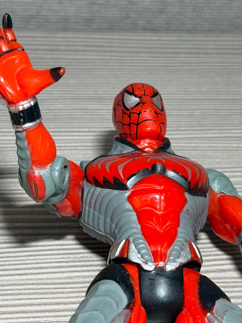 May include: A red and gray Spider-Man action figure. The figure has a red head with silver eyes and a black web design. The body is gray with red accents. The figure is posed with one arm raised.