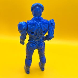 May include: A blue action figure of a humanoid character, viewed from the back, against a yellow background. The figure has detailed armor-like features and articulated joints. The figure is approximately 15 cm tall.