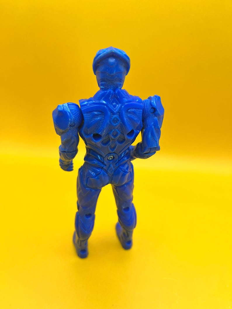 May include: A blue action figure of a humanoid character, viewed from the back, against a yellow background. The figure has detailed armor-like features and articulated joints. The figure is approximately 15 cm tall.