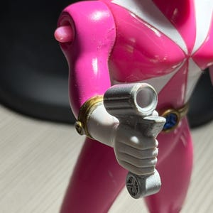 May include: A pink Power Ranger action figure with white accents and a black helmet. The figure is holding a gray weapon. The figure has gold and blue details on the belt and wrist. The figure is standing on a light-colored surface.