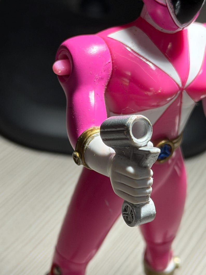 May include: A pink Power Ranger action figure with white accents and a black helmet. The figure is holding a gray weapon. The figure has gold and blue details on the belt and wrist. The figure is standing on a light-colored surface.