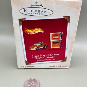 May include: A Hallmark Keepsake Ornament set featuring a red and orange Hot Wheels Sizzlers car and a red gas pump with the text "Juice Machine" and "Sizzlers". The box reads "Juice Machine™ and Revvin' Heaven Hot Wheels® Sizzlers® Set".