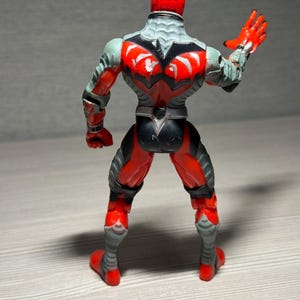 May include: A red and gray action figure with a detailed design. The figure has a red helmet, red hands, and red boots. The back of the figure features a black and red design with gray accents. The figure is posed with one arm raised.